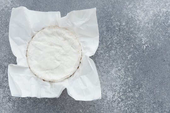 Round brie cheese wrapped in white paper on a grey surface