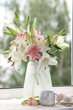 Beautiful lily flowers in vase, zephyrs and cup of drink on window sill indoors