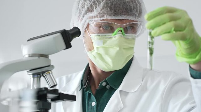 Scientific professional wearing green protective equipment examining green filled test tube under microscope, performing laboratory research with precision. Medicine, healthcare and science concept