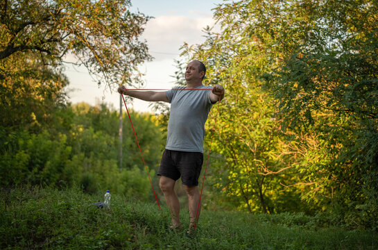 Man stretches with resistance band in forest clearing, arms extended against red tension cords. Fitness workout in natural setting with determined expression and barefoot stance on grassy ground.