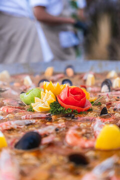 Selective-focus vertical view of carved tomato and citrus garnish arranged at the center of a large seafood paella.