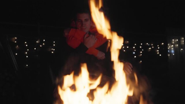 Happy boyfriend and girlfriend cuddling near warm campfire flames at campsite avoiding hypothermia risk