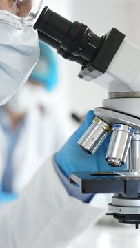 Close up of male researcher examining sample through microscope, wearing blue safety gear in modern laboratory setting. Medicine, healthcare and science concept