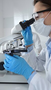 Female scientist wearing mask and blue gloves using microscope doing scientific research in modern laboratory with colleague working in background. Medicine, healthcare and science concept