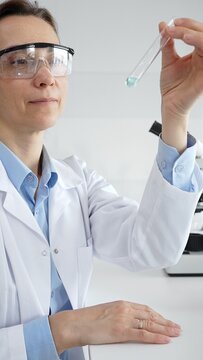 Female scientist wearing lab coat and protective glasses holding a test tube with blue liquid, conducting research in a modern laboratory setting. Medicine, health care and science concept