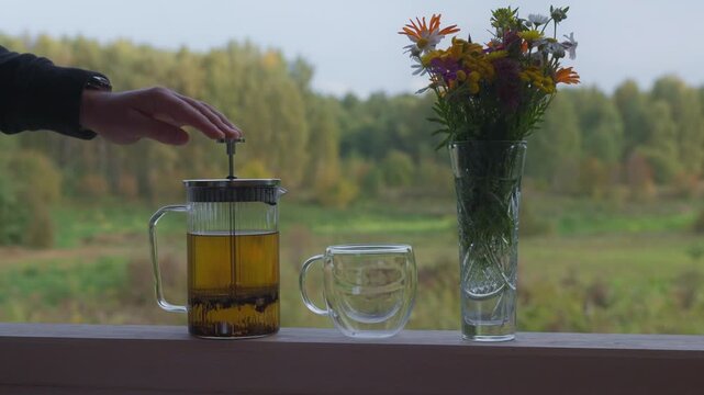 Hand prepares freshly steeped tea in glass press pot on outdoor railing. Person lifts beverage maker near flower vase and cup, showcasing cozy rustic lifestyle during golden autumn season.