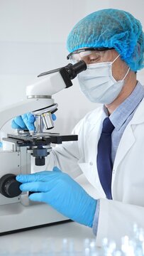 Senior scientist wearing a protective mask, cap, and gloves, examining samples under a microscope in a modern laboratory. Medicine, healthcare and science concept