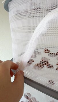 A person's hand gently holds a white mesh fabric attached to a window screen, revealing a pattern of reddish brown spots on the material, suggesting a barrier or covering in a window setting