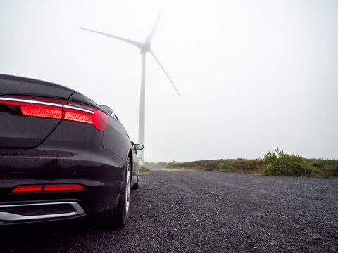 Black car drives on gravel road toward wind turbine. Misty sky creates moody atmosphere. Wind turbine stands tall in distance. Scene blends modern technology with natural landscape
