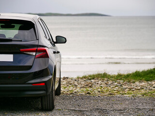 Black car parked on rocky beach beside calm sea. Gentle waves lap shore near distant island. Scene feels serene, peaceful, and solitary. Ideal for travel, vacation, or coastal lifestyle themes © mark_gusev
