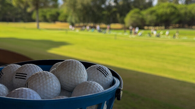 Basket of used golf balls at a driving range with green golf course and players blurred in the background, outdoor sports practice and training concept.
