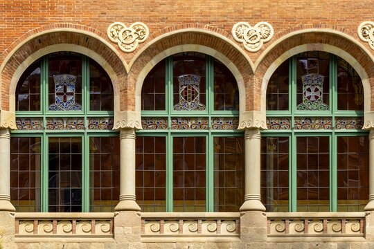Three arched windows featuring intricate stained glass crests and decorative pillars against a brick facade at Hospital de Sant Pau in Barcelona. Showcasing distinctive modernisme architectural style