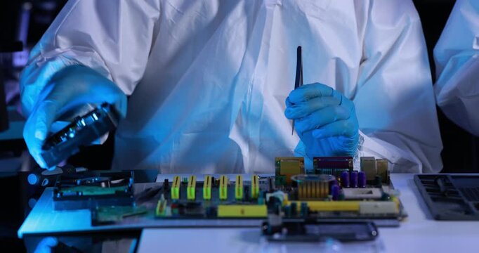Close-up of a technician in protective clothing repairing an electronic circuit board with a screwdriver under blue laboratory light.