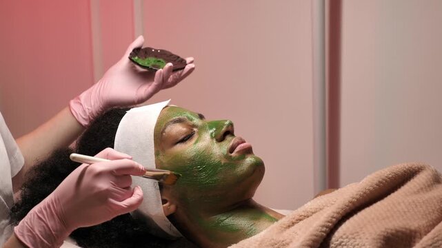 Cosmetologist applying green clay face mask with brush on female client face in spa salon. Skin care treatment, organic cosmetic facial procedure, rejuvenation, facial care and wellness concept