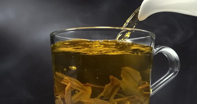 Hot green tea pouring into glass cup with tea leaves and steam in slow motion
