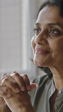 Vertical video: Sighing, Indian woman holding clasped hands, reflecting by window with bindi