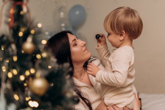 Mother holding toddler beside glowing Christmas tree at home sharing tender eye contact festive family bonding authentic parenting holiday tradition and warm childhood memories in cozy winter season