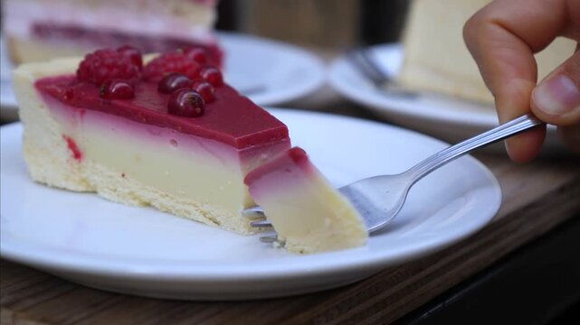Fork separates cheesecake bite from raspberry topped slice on white plate. Hand holds utensil steady while creamy filling and crumb base stay visible, creating fresh dessert serving moment.