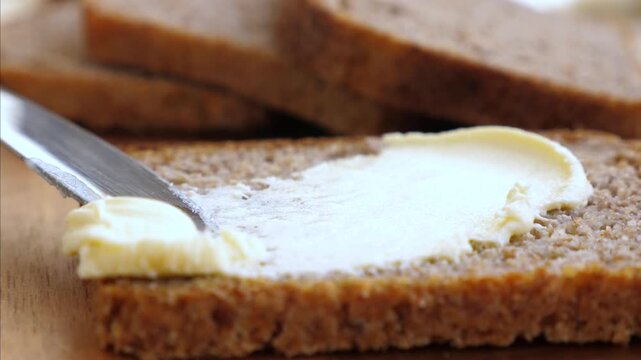 Butter knife spreads soft butter on sliced bread, slowly smoothing one piece in close up kitchen prep. Warm toasted loaves sit behind, showing simple breakfast routine with calm focus.