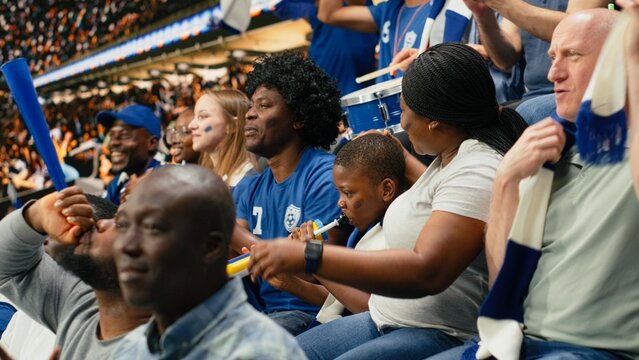 Young soccer fan cheering with blue supporters during live football match in packed stadium filled with cheering supporters; family outing captures fatherhood and role model during soccer experience.