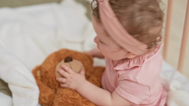 Baby girl in pink onesie holding colorful rattle, chewing teether and touching teddy with hand inside building crib with soft blanket; curiosity.