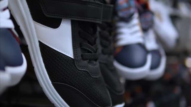 Black and white sneakers swing in tight closeup, with other shoes softly out of focus behind. Hanging footwear display presents shoes swaying together, with visible laces, soles, and textured uppers.