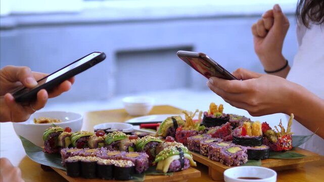 Group shares sushi lunch while holding smartphones over wooden table. Colorful maki and nigiri sit beside sauce bowls, and relaxed conversation feels casual, contemporary and pleasantly connected.