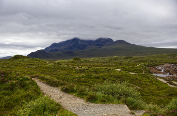 Fototapeta premium Hiking trail leading through green moorland towards the dramatic dark peaks of the Black Cuillin mountains on the Isle of Skye, Scotland.