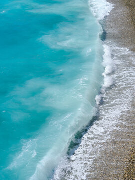 Vertical photo. Big storm waves crashing on a pebble beach with azure water. Summer sea coastline during powerful ocean weather.
