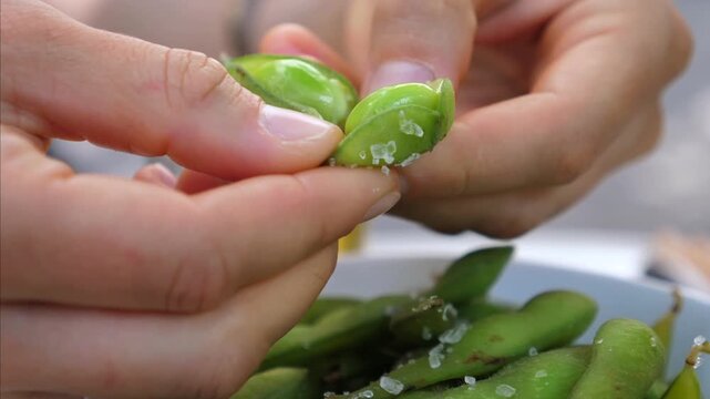 Hands shell edamame in close up. Salted green pods are pinched open over white bowl, revealing bright beans while calm meal moment unfolds at table. Fingers separate pod halves for serving.