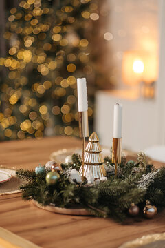 Christmas table centerpiece with candles, wreath, and golden tree ornament against blurred christmas tree lights.