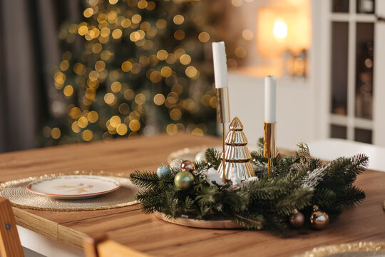 Christmas table centerpiece with candles, wreath, and golden tree ornament against blurred christmas tree lights.