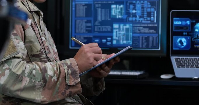 Military service member in camouflage writes on a clipboard beside computer screens technical data in a dark operations room.