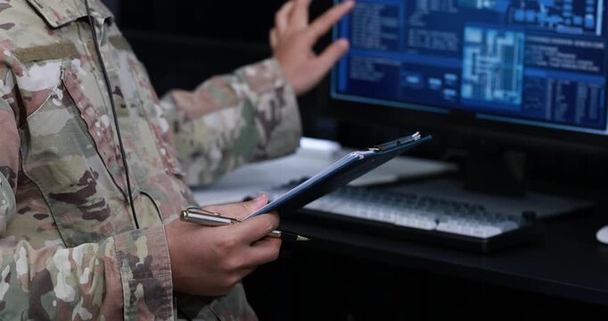 Military service member in camouflage uniform holds a clipboard and pen while monitoring digital information on a computer screen in a dark operations room.