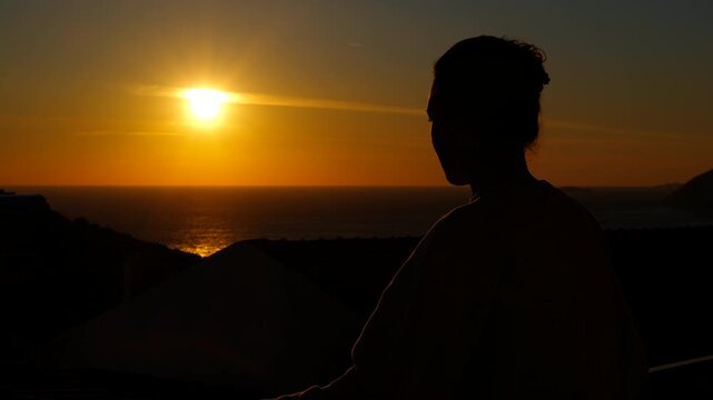 Woman watching the sunset over the ocean. Silhouette of a pensive woman looking at the orange sun setting over the calm sea, a serene moment, with a beautiful and scenic view
