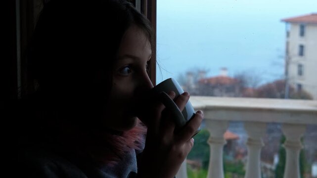 Young girl enjoying a hot beverage on a balcony with a scenic view. Backlit silhouette of young girl enjoying peaceful moment, sipping warm drink while gazing through balcony window