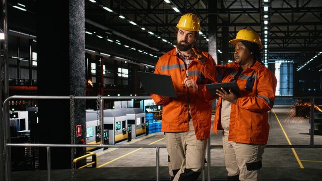 Diverse engineers monitor heavy equipment in an industrial plant while metallic frameworks and machinery define the production line and engineering facility.