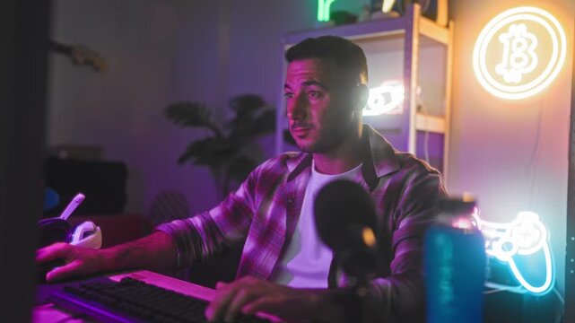 Man raising arms over keyboard with headset nearby and neon controller sign on shelf in gaming studio celebrating a win; excitement victory.