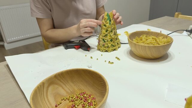 Hands decorate pasta Christmas tree with small pearl beads and festive details. DIY holiday craft for school or kindergarten. Good for seasonal tutorials, family creativity, handmade decor and