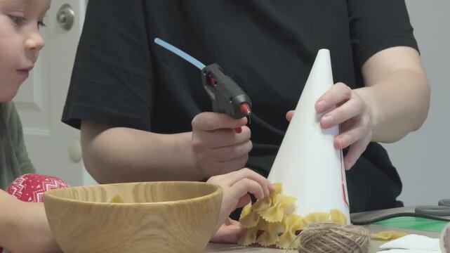 Mother and child creating Christmas craft using pasta bow shapes. DIY decoration for school or kindergarten.