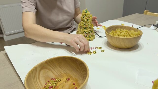 Woman creates handmade Christmas tree craft from bow tie pasta at table. Holiday DIY activity for school or kindergarten. Suitable for parenting, education, seasonal creativity, handmade decor