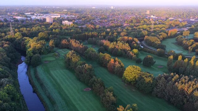 Aerial video tracking the blue River Mersey as it meanders alongside green fairways, sand bunkers, and mature woodland near East Didsbury, Manchester at dawn.