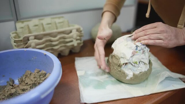 Hands shaping papier mache over rounded form on tabletop in indoor craft workspace with blue clay bowl nearby, showing careful handmade recycling movement