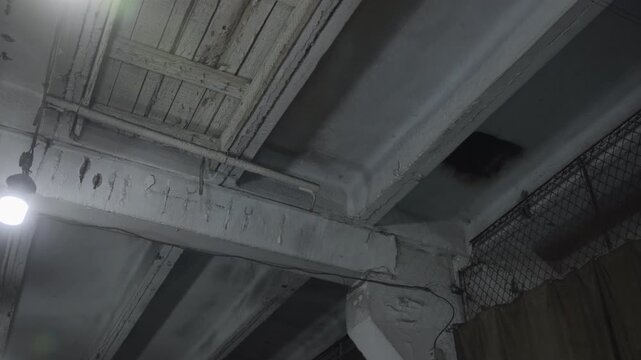Worn concrete ceiling with exposed beams, cracked paint, hanging lamp, pipes, and dark recesses in cool gray industrial interior with rough texture, diagonal structure, and neglected urban mood
