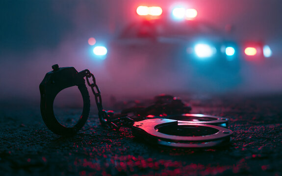 Police raid at night and you are under arrest concept. Silhouette of handcuffs with police car on backside with the flashing red and blue police lights at foggy background