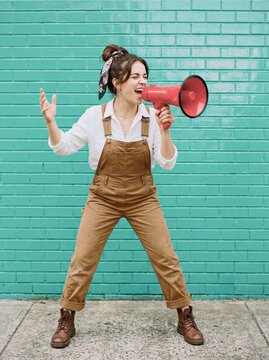 Woman in brown overalls shouting into red megaphone at urban rally against blue brick wall