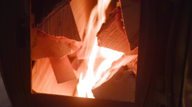 Bright flames rising inside wood burning stove with split logs, glowing embers, rough timber grain, dark metal surround, warm orange light, strong contrast, rustic cozy heat and winter comfort mood