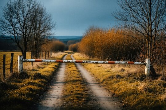 Old weathered barrier blocks rural dirt road. Orange, white metal gate closes access to countryside lane through autumn field. Quiet landscape with trees, shrubs under cloudy sky. Remote path leads