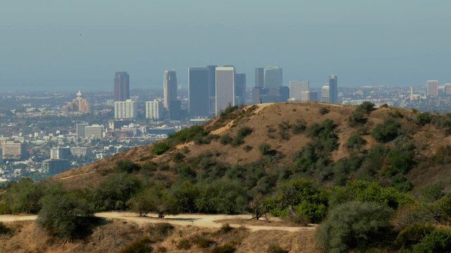 Hikers on a dirt trail across dry hills with the Century City skyline behind. Los Angeles, California on a hazy day. Green shrubs, golden grass and a lone figure on the hilltop. Telephoto view.