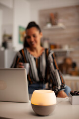 Fototapeta premium Woman works at a laptop while a glowing aromatherapy diffuser mists in the foreground, combining remote productivity with a calm home-wellness environment, supporting remote work wellness.